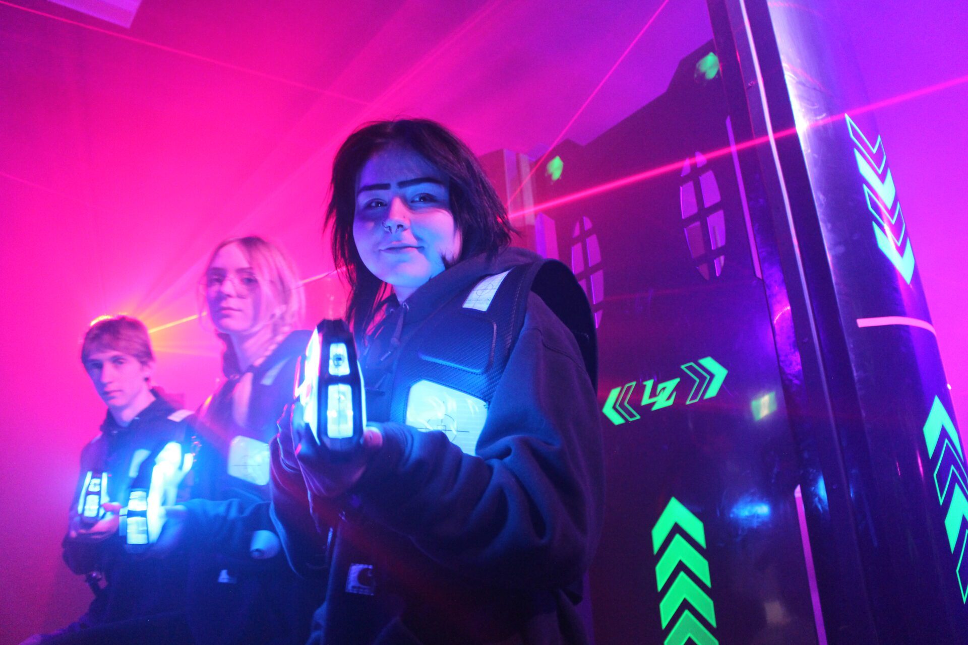Three teenagers - a boy and two girls - stand in a staggered line next to a black wall with flourescent yellow arrows painted on it. The atmosphere is smokey and there are red laser beams cutting through the background. There are pink, purple, and blue lights colouring the smoke and the faces of the teens. All three hold laser tag guns - about the length of their forearms and glowing with blue light. They point their guns at the camera. The two teens in the background have serious expressions on their faces. The teen in the foreground - a girl - has a small smile on her face as the looks straight into the camera and points her gun.